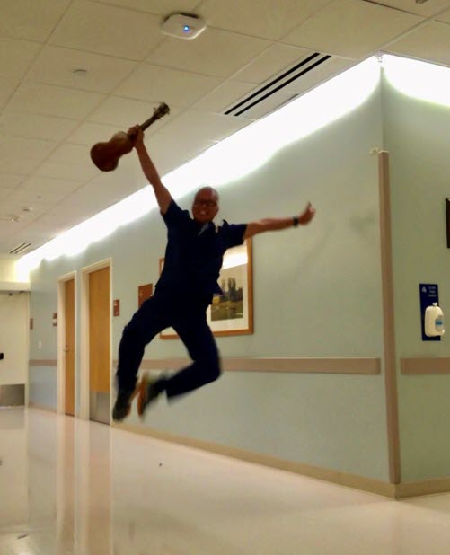 Travel nurse, Rod, leaps in a hospital hallway with his ukulele in hand. 